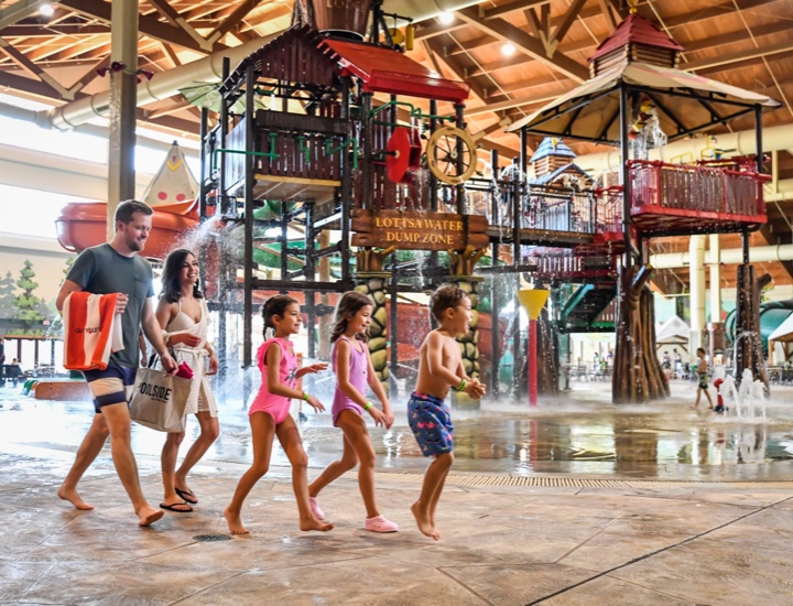 Family wearing swim gear walks in front of a splash pad structure inside a pavilion
