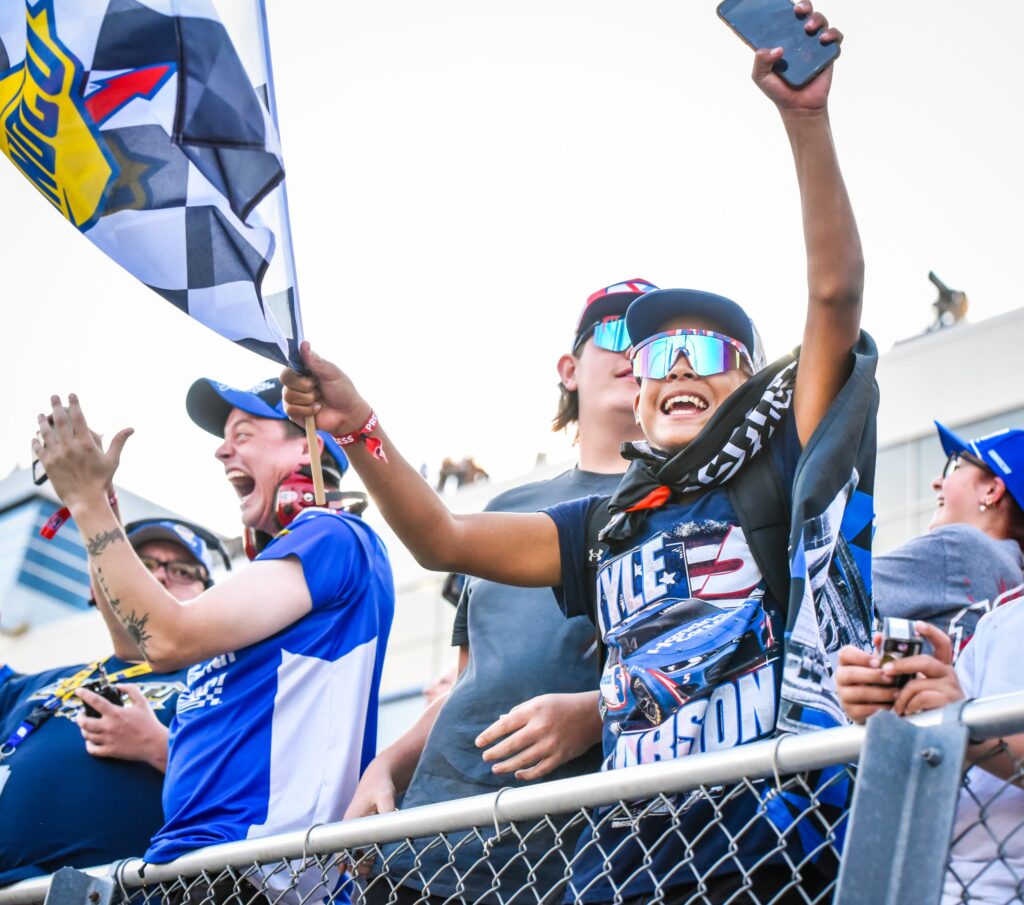 Enthusiastic racing fans clap and wave flags behind a low fence