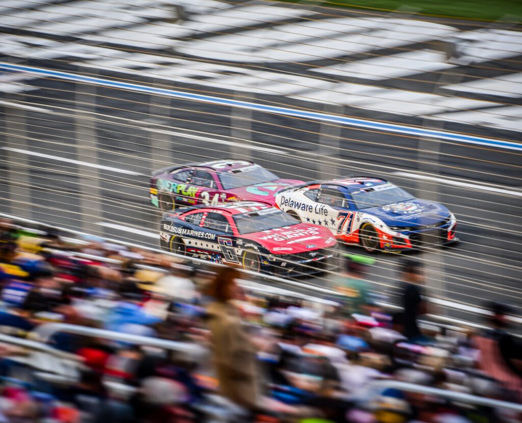 Blurred image of three cars racing on a track, with a large audience in the stands