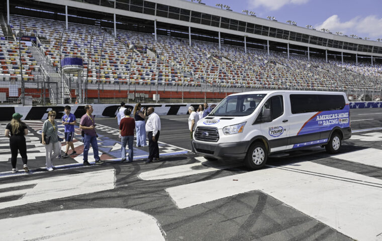 Photograph of a group of people standing on a racetrack near a white shuttle van with "America's Home For Racing" branding. The scene includes empty grandstands with multicolored seats and clear blue sky, indicating a motorsport event or tour setting.