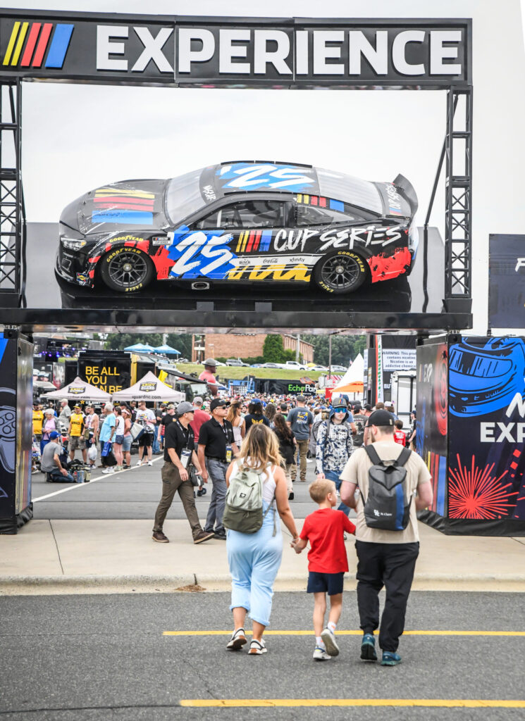 Photograph of a NASCAR-themed event entrance featuring a large display of a black race car with blue and red accents and "25 Cup Series" branding mounted above a walkway. A family of three walks toward a crowded outdoor event area under a sign reading "EXPERIENCE" with colorful racing stripes, highlighting fan engagement and motorsport culture.