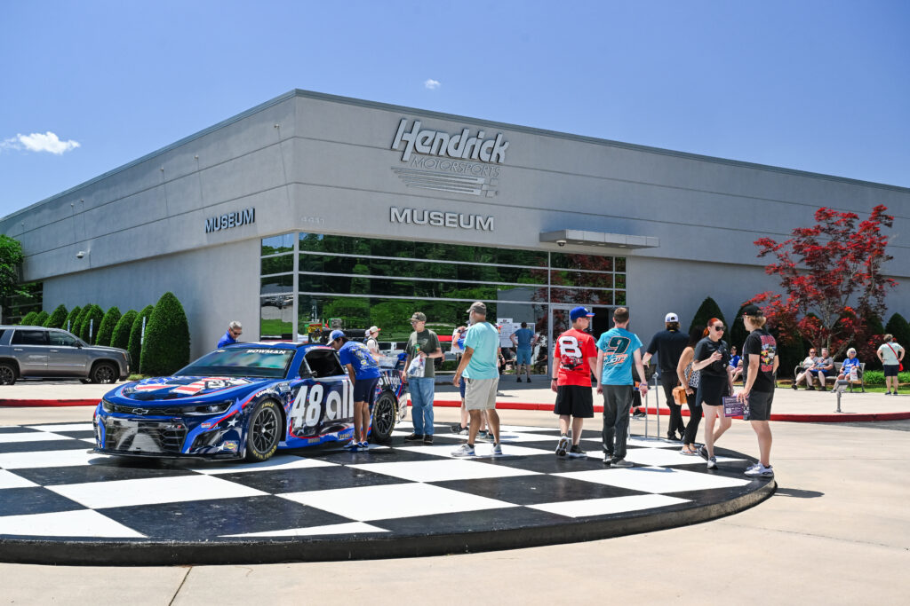 Photograph of Hendrick Motorsports Museum exterior featuring a blue and black race car with number 48 parked on a large checkered circle in front. Several people, including families and children, are walking and standing around the car and museum entrance.