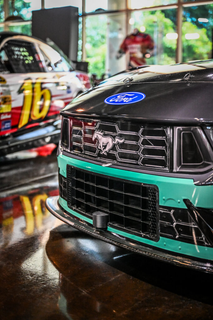 The image shows two Ford Mustang race cars parked in an indoor showroom with a polished, reflective floor.
